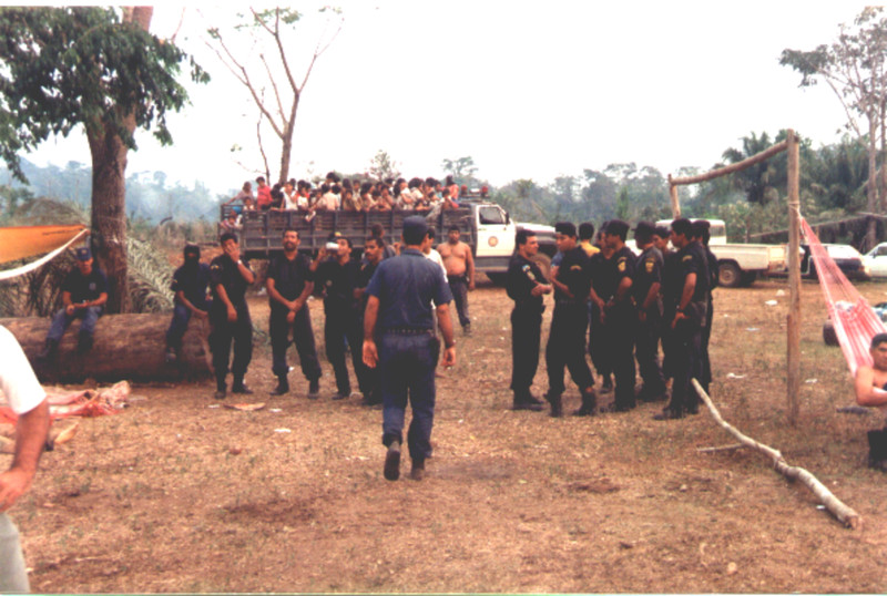 Após cometerem todo tipo de atrocidade, policiais conversam, bebem e descansam tranquilamente ao lado dos corpos de camponeses assassinados (lado esquerdo). No fundo camponeses prisioneiros em cima de caminhão.